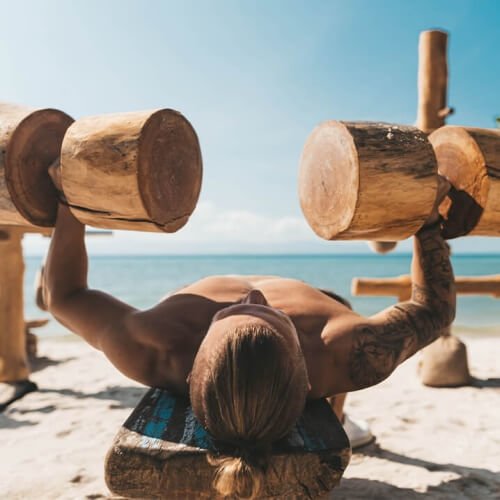 Mann trainiert mit Hanteln aus Holz am Strand mit blauem Meer